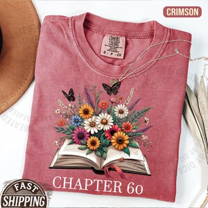 May include: A crimson short-sleeve t-shirt featuring a graphic of an open book with a colorful bouquet of flowers and two black butterflies emerging from the pages. The text "CHAPTER 60" is printed in white below the book. A Comfort Colors label is visible.