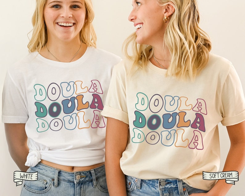 May include: Two women wearing white and cream t-shirts with the word "Doula" repeated in a retro font style. The shirts are available in white and soft cream.