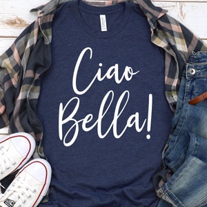 K&ouml;nnte beinhalten: Marineblaues T-Shirt mit dem wei&szlig;en Schriftzug "Ciao Bella!". Das Shirt wird mit einer blauen Jeans, einem karierten Hemd und wei&szlig;en Turnschuhen kombiniert. Eine Uhr mit braunem Lederarmband ist ebenfalls zu sehen.