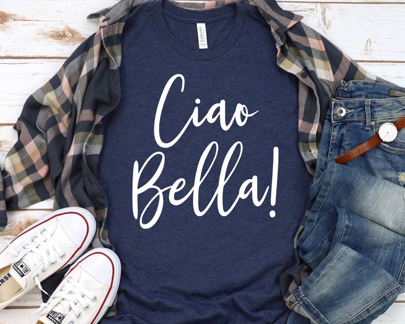 K&ouml;nnte beinhalten: Marineblaues T-Shirt mit dem wei&szlig;en Schriftzug "Ciao Bella!". Das Shirt wird mit einer blauen Jeans, einem karierten Hemd und wei&szlig;en Turnschuhen kombiniert. Eine Uhr mit braunem Lederarmband ist ebenfalls zu sehen.