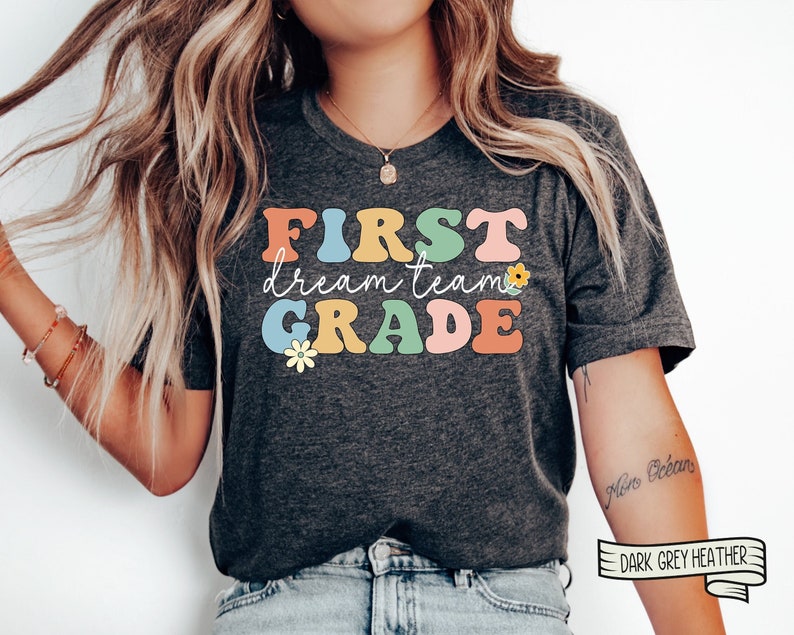 Op de afbeelding: Een donkergrijs heather t-shirt met de tekst "FIRST dream team GRADE" in kleurrijke retro letters en een kleine bloem grafiek.