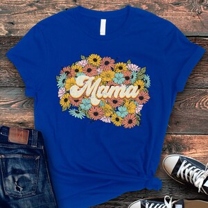 Puede incluir: Una camiseta azul real con un dise&ntilde;o floral de estilo retro y la palabra "Mama" en una fuente amarilla, naranja y marr&oacute;n.