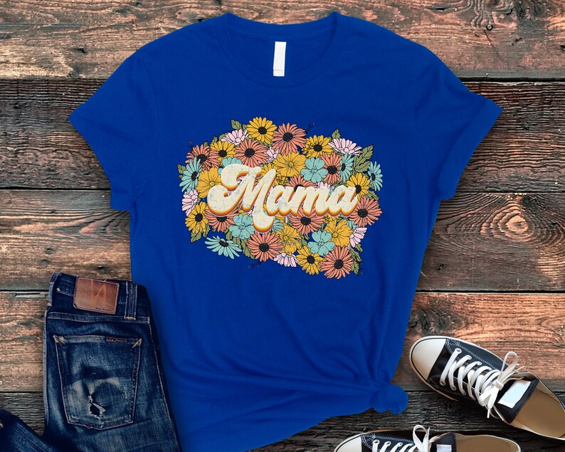 Puede incluir: Una camiseta azul real con un dise&ntilde;o floral de estilo retro y la palabra "Mama" en una fuente amarilla, naranja y marr&oacute;n.