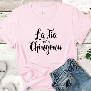 Może przedstawiać: Jasnor&oacute;żowa koszulka z czarnym napisem "La Tia m&aacute;s Chingona". Na zdjęciu znajdują się dżinsowe szorty, czerwone okulary i srebrne sandały, wszystko na drewnianej powierzchni.