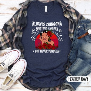 May include: A navy blue t-shirt with a graphic of a woman with red roses and the text "Always Chingona Sometimes Cabrona - But Never Pendeja -" in white. The t-shirt is on a wooden surface with a plaid shirt, jeans, and white sneakers.