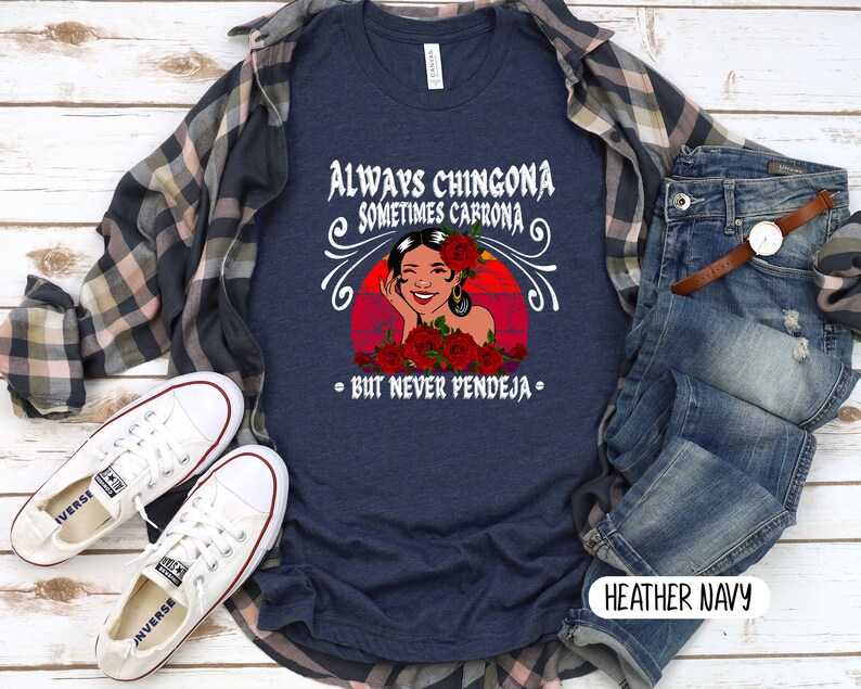 May include: A navy blue t-shirt with a graphic of a woman with red roses and the text "Always Chingona Sometimes Cabrona - But Never Pendeja -" in white. The t-shirt is on a wooden surface with a plaid shirt, jeans, and white sneakers.