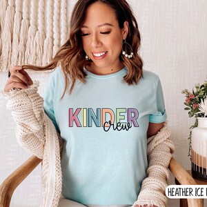 May include: A light blue t-shirt with the text "Kinder Crew" in rainbow colors. The shirt is being worn by a person with long brown hair and is paired with a cream cardigan.