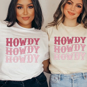 May include: Two women wearing white and cream t-shirts with the word "HOWDY" printed in pink and red. The word is stacked vertically four times on each shirt.
