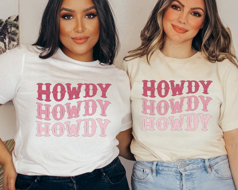 May include: Two women wearing white and cream t-shirts with the word "HOWDY" printed in pink and red. The word is stacked vertically four times on each shirt.