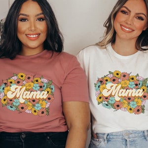 Puede incluir: Dos mujeres con camisetas con un dise&ntilde;o floral y la palabra "Mama" en una fuente retro. Las camisetas son de diferentes colores, una es rosa y la otra es blanca.