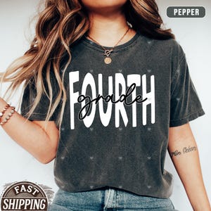 Op de afbeelding: Een grijs T-shirt met de tekst "FOURTH grade" in witte letters. Het shirt wordt gedragen door een persoon met lang bruin haar.