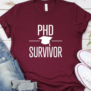 May include: Burgundy t-shirt with white text that reads "PhD Survivor" and a graduation cap graphic.