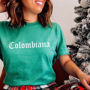 Op de afbeelding: Een groen T-shirt met het woord "Colombiana" in wit op de voorkant. Het shirt wordt gedragen door een persoon in rood-zwarte geruite pyjamabroek.