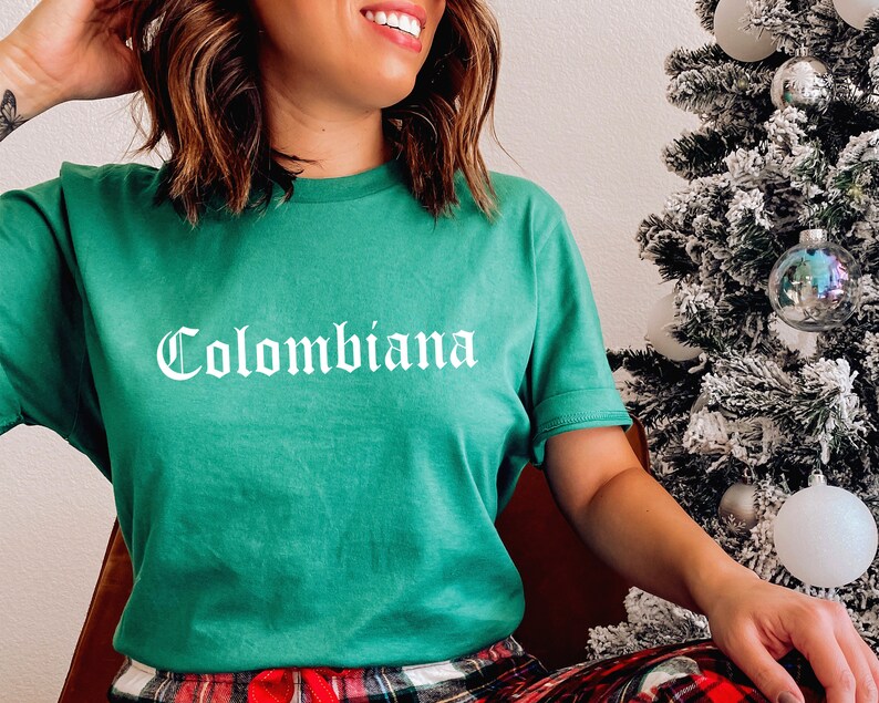 Op de afbeelding: Een groen T-shirt met het woord "Colombiana" in wit op de voorkant. Het shirt wordt gedragen door een persoon in rood-zwarte geruite pyjamabroek.
