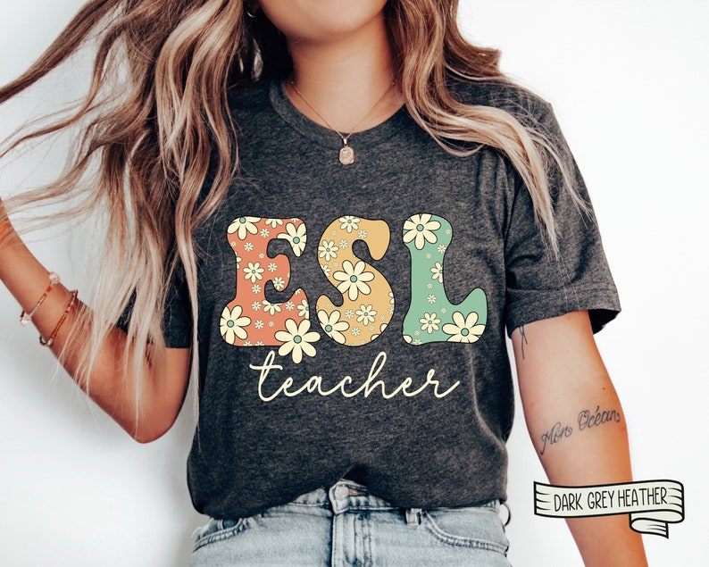 May include: A dark grey heather t-shirt with the text "ESL teacher" in a retro font with floral accents. The letters are outlined in yellow and filled with orange, yellow, and green floral patterns.