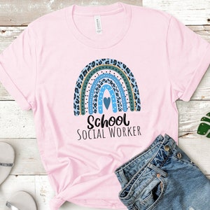 Puede incluir: Camiseta rosa con un dise&ntilde;o de arco&iacute;ris azul, verde y dorado y el texto "School Social Worker".