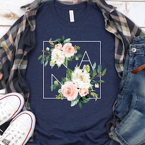 May include: A navy blue t-shirt with a white square frame around a floral design. The design features pink and white flowers with green leaves. The word "NANA" is spelled out in the frame.