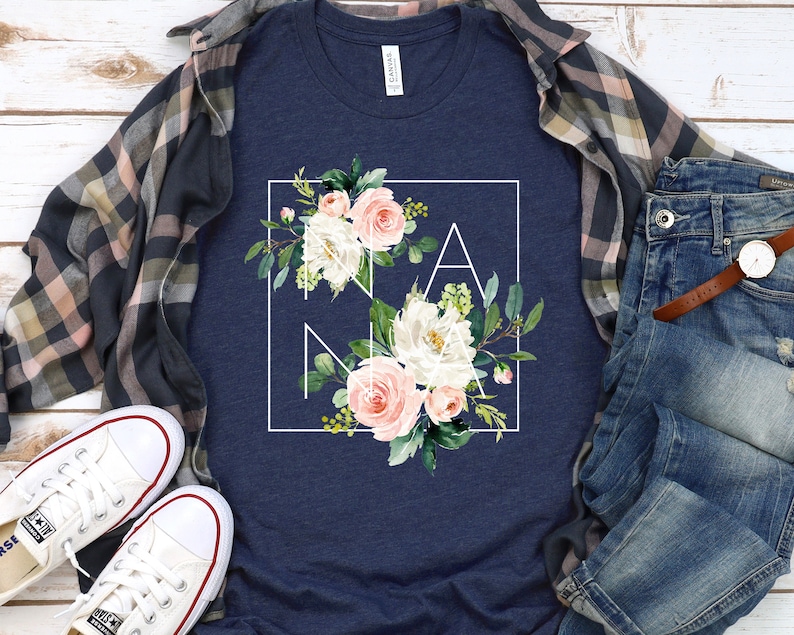 May include: A navy blue t-shirt with a white square frame around a floral design. The design features pink and white flowers with green leaves. The word "NANA" is spelled out in the frame.