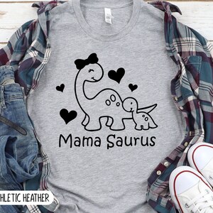 May include: A heather grey t-shirt with a black and white graphic of a mama dinosaur with a bow and a baby dinosaur. The text "Mama Saurus" is printed below the dinosaurs.