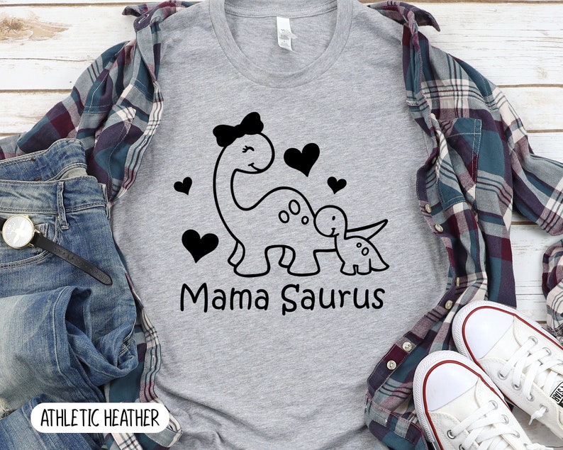May include: A heather grey t-shirt with a black and white graphic of a mama dinosaur with a bow and a baby dinosaur. The text "Mama Saurus" is printed below the dinosaurs.