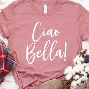 K&ouml;nnte beinhalten: Ein staubrosafarbenes T-Shirt mit dem Schriftzug "Ciao Bella!" in wei&szlig;er Schrift. Das Shirt ist mit einem karierten Schal, Jeans und einem Paar beiger Schuhe gestylt. Das Shirt ist aus weichem Material.