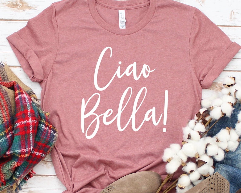K&ouml;nnte beinhalten: Ein staubrosafarbenes T-Shirt mit dem Schriftzug "Ciao Bella!" in wei&szlig;er Schrift. Das Shirt ist mit einem karierten Schal, Jeans und einem Paar beiger Schuhe gestylt. Das Shirt ist aus weichem Material.