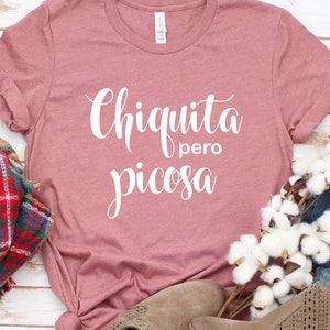 May include: A dusty rose colored t-shirt with the text "Chiquita pero picosa" in white cursive lettering.