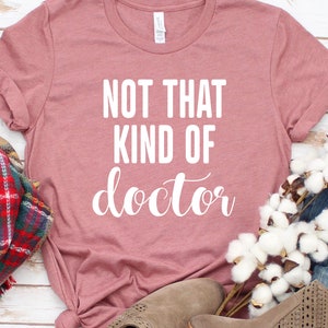 May include: A dusty rose colored t-shirt with the text "NOT THAT KIND OF doctor" in white lettering.