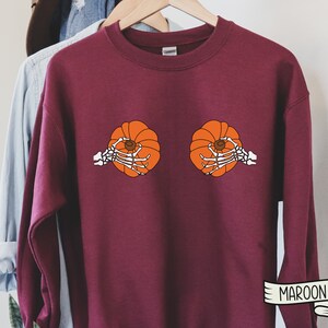 Op de afbeelding: Een maroon sweatshirt met een afbeelding van twee pompoenen die door skelethandjes worden vastgehouden. De tekst "MAROON" is gedrukt op een wit lint in de rechterbenedenhoek.
