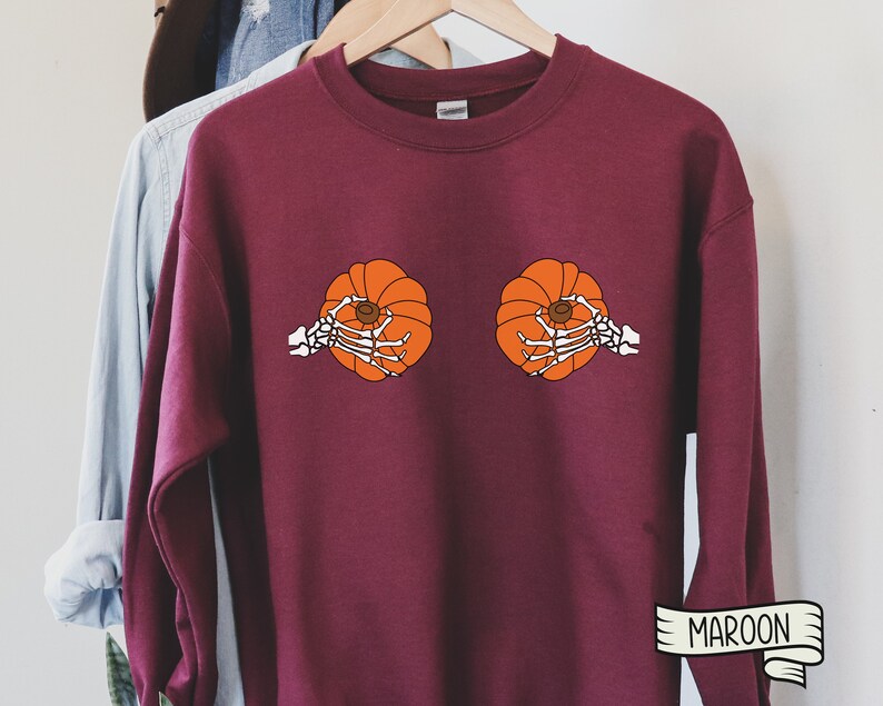 Op de afbeelding: Een maroon sweatshirt met een afbeelding van twee pompoenen die door skelethandjes worden vastgehouden. De tekst "MAROON" is gedrukt op een wit lint in de rechterbenedenhoek.