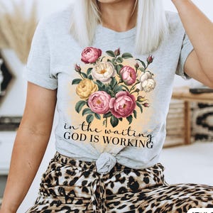 May include: A light gray t-shirt featuring a floral design with pink, yellow, and white roses. The text "in the waiting GOD IS WORKING" is printed below the flowers. The shirt is knotted at the waist.