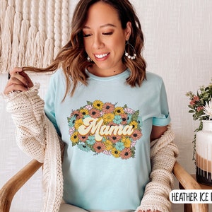 Puede incluir: Una camiseta azul claro con un dise&ntilde;o floral y la palabra "Mama" en una fuente retro. La camiseta est&aacute; hecha de un material suave y c&oacute;modo y es perfecta para el uso diario.