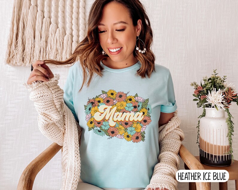 Puede incluir: Una camiseta azul claro con un dise&ntilde;o floral y la palabra "Mama" en una fuente retro. La camiseta est&aacute; hecha de un material suave y c&oacute;modo y es perfecta para el uso diario.
