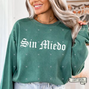 May include: A light green sweatshirt with the phrase "Sin Miedo" in white gothic lettering. The sweatshirt has a crew neck and long sleeves. Small heart-shaped patterns are scattered across the fabric. The words "Light Green" are written on a banner.