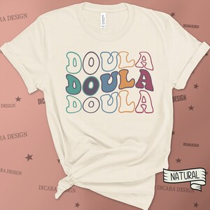May include: A beige t-shirt with the word "DOULA" written three times in a retro font with colorful outlines. The word is stacked vertically with each word in a different color.