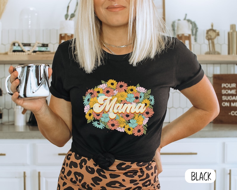 May include: A black t-shirt with a floral design and the word "Mama" in a retro font. The shirt is being worn by a person with blonde hair.