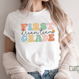 Op de afbeelding: Wit T-shirt met de tekst "FIRST dream team GRADE" in kleurrijke retro-letters en een kleine bloemgrafiek.