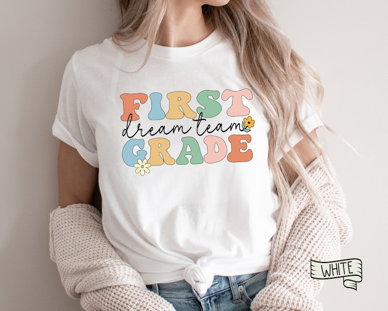 Op de afbeelding: Wit T-shirt met de tekst "FIRST dream team GRADE" in kleurrijke retro-letters en een kleine bloemgrafiek.