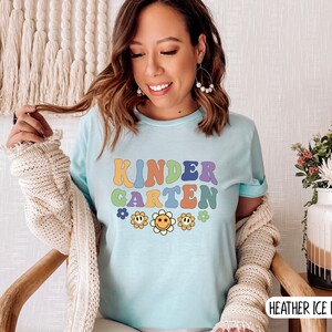 Puede incluir: Una camiseta azul claro con el texto "KINDERGARTEN" en letras retro de colores. El texto est&aacute; rodeado de peque&ntilde;as flores amarillas con caras sonrientes.