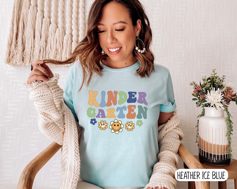 Puede incluir: Una camiseta azul claro con el texto "KINDERGARTEN" en letras retro de colores. El texto est&aacute; rodeado de peque&ntilde;as flores amarillas con caras sonrientes.
