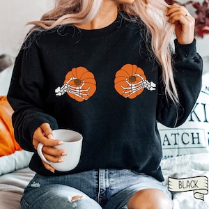 Op de afbeelding: Zwart sweatshirt met een afbeelding van twee oranje pompoenen met skelethandjes die ze vasthouden. De tekst "BLACK" is zichtbaar in de rechterbenedenhoek van de afbeelding.