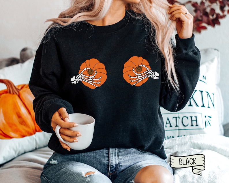Op de afbeelding: Zwart sweatshirt met een afbeelding van twee oranje pompoenen met skelethandjes die ze vasthouden. De tekst "BLACK" is zichtbaar in de rechterbenedenhoek van de afbeelding.