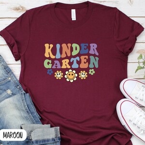 Puede incluir: Camiseta color granate con el texto "KINDERGARTEN" en una fuente retro arco&iacute;ris con gr&aacute;ficos de flores sonrientes.