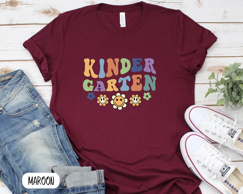 Puede incluir: Camiseta color granate con el texto "KINDERGARTEN" en una fuente retro arco&iacute;ris con gr&aacute;ficos de flores sonrientes.