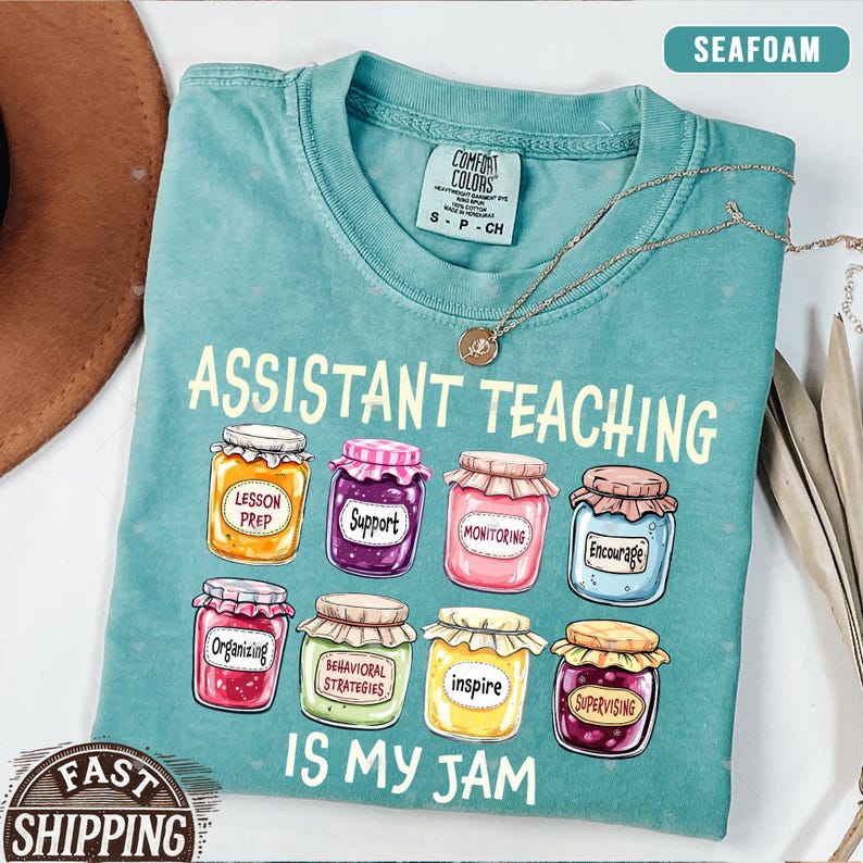 Puede incluir: Una camiseta azul turquesa con el texto "Assistant Teaching is my jam" en amarillo. El texto est&aacute; rodeado de tarros coloridos con diferentes palabras en ellos, como "Lesson Prep", "Support", "Monitoring", "Encourage", "Organizing", "Behavioral Strategies", "Inspire" y "Supervising".