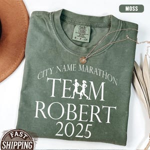 May include: A moss green t-shirt with the text "CITY NAME MARATHON TEAM ROBERT 2025" in white. The shirt has a crew neck and short sleeves. A gold necklace with a pendant is draped across the shirt.