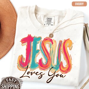 May include: Ivory-colored t-shirt with the words "JESUS Loves You" in a colorful, brushstroke design. The shirt has a crew neck and short sleeves. A gold necklace with a pendant is draped on the shirt.