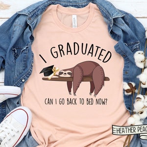 May include: A peach-colored t-shirt with the text "I Graduated Can I Go Back To Bed Now?" and a cartoon sloth wearing a graduation cap. The sloth is lying on a branch with a diploma.