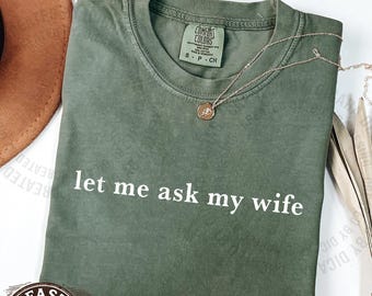 May include: A moss green t-shirt with the text "let me ask my wife" in white. The shirt has a crew neck and short sleeves. A gold necklace with a pendant is draped across the shirt.