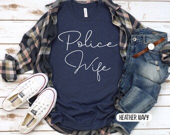 May include: A navy blue t-shirt with the text "Police Wife" in white script. The shirt is laid flat on a white wooden surface with a plaid shirt, jeans, and white sneakers.